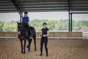 A rider in equestrian attire demonstrates correct dressage alignment while mounted on a black gelding exhibiting an uphill frame, as another individual stands nearby smiling. Both are inside a covered arena with a backdrop of trees visible through the open sides.