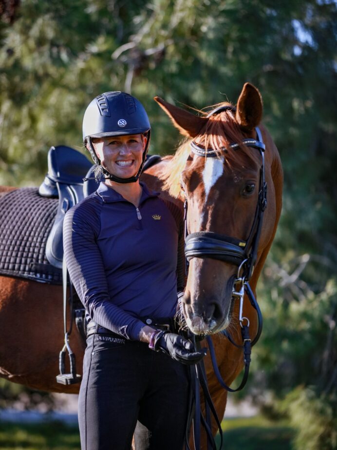 A smiling person in a riding helmet and gear stands outdoors, holding the reins of a brown horse with a black saddle, ready for Dressage Training against a backdrop of green trees.