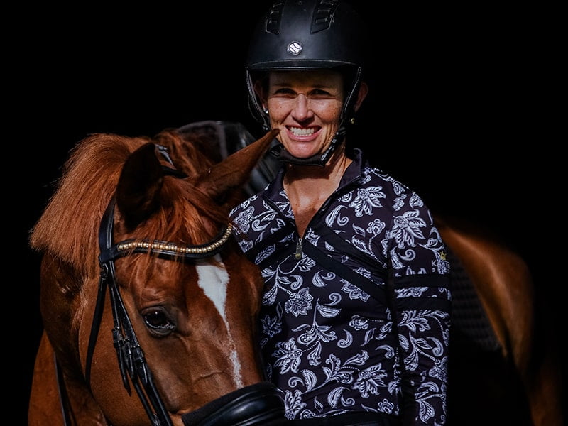A smiling person in a patterned shirt and helmet stands beside a brown horse with a white blaze, both facing the camera against a dark background.