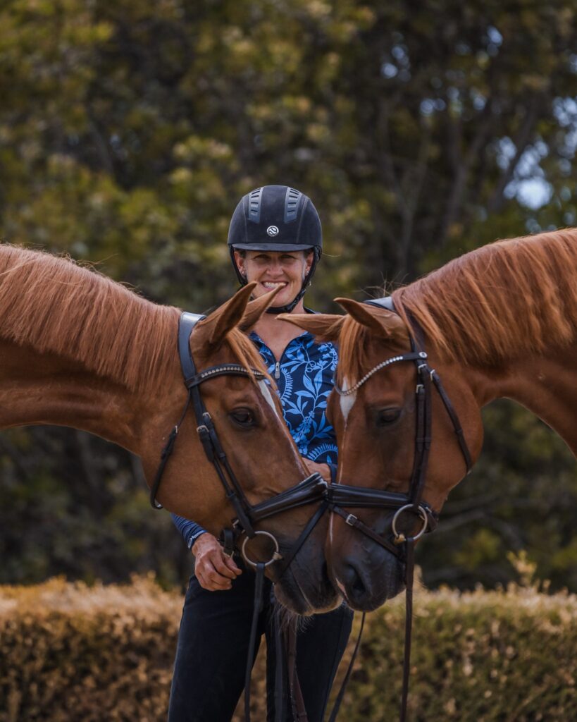 A smiling person wearing a riding helmet stands outdoors between two chestnut horses whose heads are gently touching. The background features green foliage and trimmed hedges.