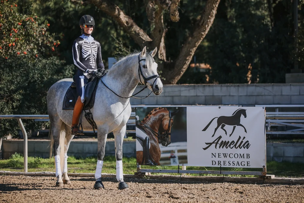 A person in a skeleton costume and helmet demonstrates excellent rider position on a gray horse near an Amelia Newcomb Dressage sign in an outdoor 2023 arena, with trees in the background and Rider Rankings on display.