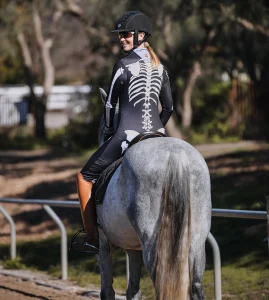 A person wearing a black bodysuit with a white skeleton print and a helmet is riding a gray horse outdoors, smiling and looking back over their shoulder, showcasing their Rider Ranking for 2023.