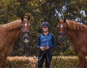 A person, featured in EuroDressage, wears a helmet and blue patterned shirt while smiling outdoors, holding the reins of two brown horses on either side. Trees and bushes fill the background.