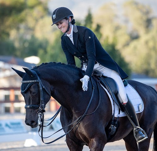 A smiling equestrian in formal riding attire pats a dark horse’s neck during a dressage event, with competition courses stretching out amid blurred greenery and distant buildings in the background.