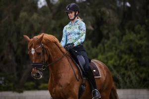A person wearing a helmet and a colorful, floral-patterned shirt rides a chestnut horse with a white marking on its forehead, practicing skills from the Training Scale Masterclass outdoors among the trees.