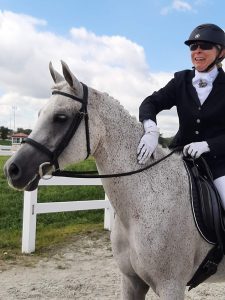 Strides Member Diana, in formal riding attire, smiles while sitting on a dapple grey horse near a white fence, with a grassy area and cloudy sky in the background.