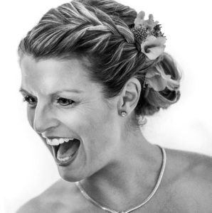 Sandra Künkel, with braided hair decorated with flowers, smiles widely and looks joyful. She wears a necklace and stud earrings. The black and white photo features a soft background.