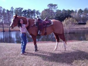 Donna, a Strides member, stands on the grass hugging the neck of a saddled brown horse near a pond, with trees and blue sky in the background.