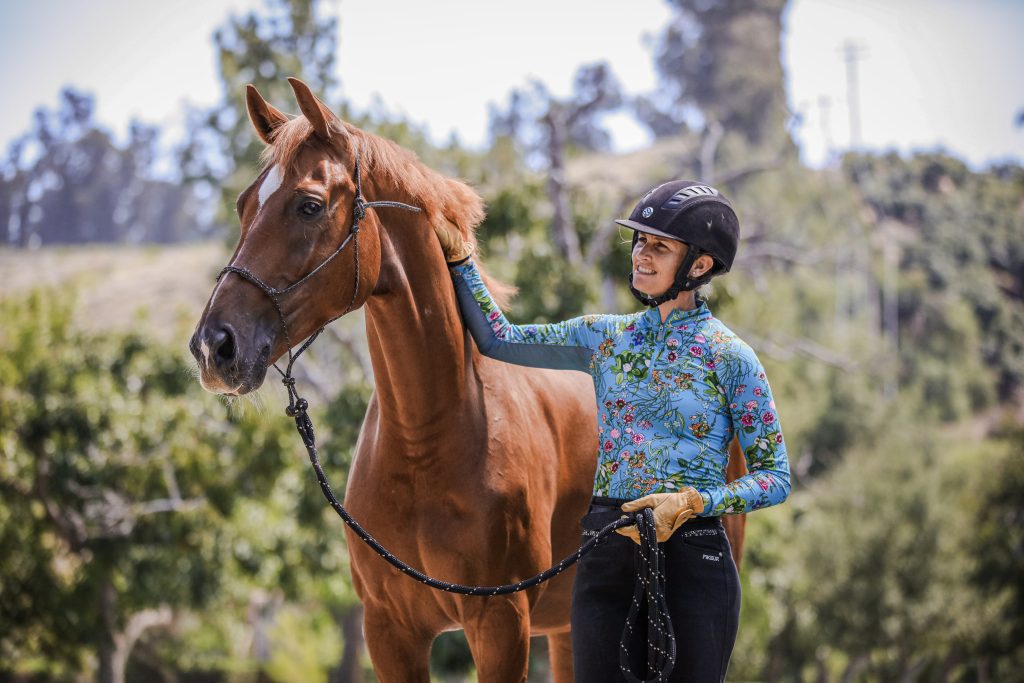 A woman in a helmet and floral long-sleeve shirt smiles while holding the bridle of a chestnut horse outdoors on a sunny day, enjoying dressage training amid green trees and blurred hills in the background.