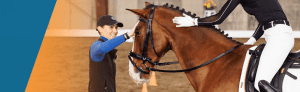 A woman in a blue shirt and black cap smiles while touching a brown horse ridden by another person during dressage training, in an indoor arena with a blurred background.