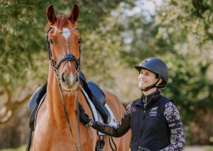 A smiling person in equestrian gear and helmet stands beside a brown horse with bridle and saddle, outdoors among green trees—perfect for the November 2023 newsletter cover.