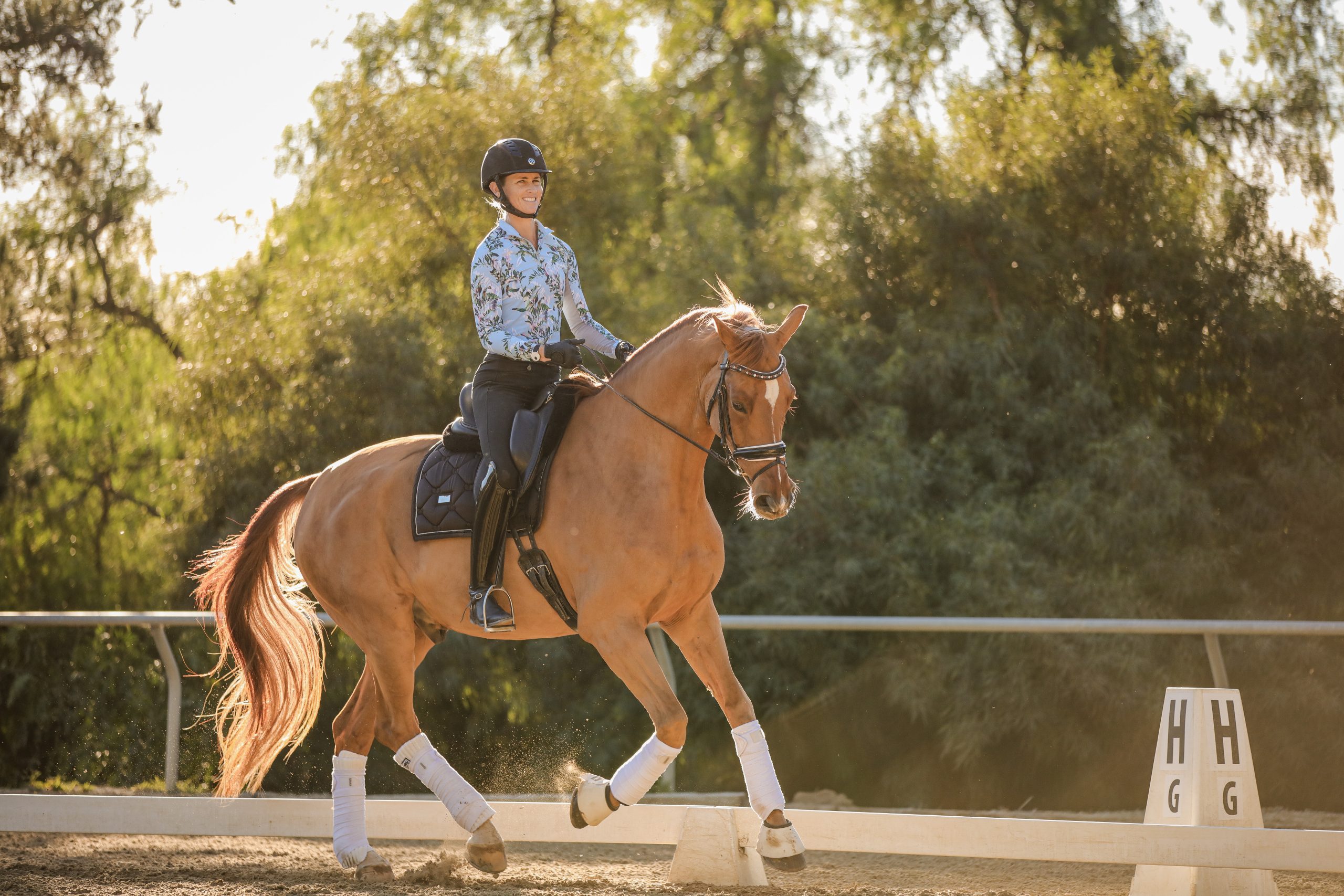 A rider in a helmet and floral shirt trots a chestnut horse with white leg wraps and a black saddle in an outdoor dressage arena, sunlight and trees in the background—perfect for listening to Heels Down Happy Hour Podcast Episode 122.
