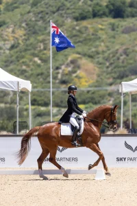 An equestrian rider in formal attire guides a chestnut horse through a Training Level dressage event, with an Australian flag and white tents in the background.