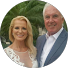 Juliette Matis A smiling woman in a white dress stands next to a smiling man in a blue suit, perhaps celebrating after competition coaching. Green palm tree leaves are visible in the blurred background.