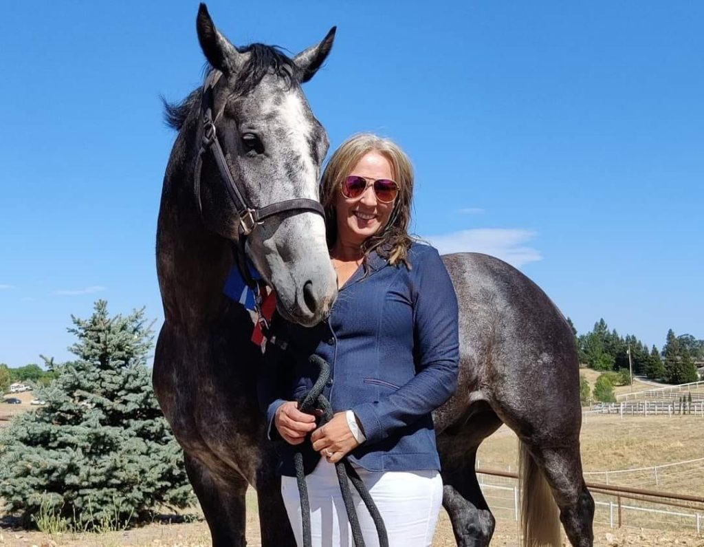 A smiling woman in sunglasses stands next to a gray horse wearing a bridle and colorful ribbon. They're outdoors on a sunny day, enjoying the rural landscape—ready for their 2024 Riding Bootcamp adventure beneath the pine tree.