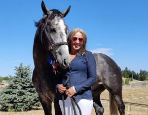 A smiling woman in sunglasses stands next to a gray horse wearing a bridle and colorful ribbon. They're outdoors on a sunny day, enjoying the rural landscape—ready for their 2024 Riding Bootcamp adventure beneath the pine tree.