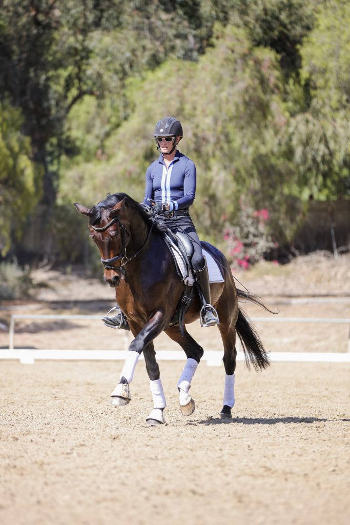 A person in a navy blue riding outfit and helmet is riding a dark brown horse with white leg wraps in an outdoor arena, surrounded by trees and sunlight—perfect for building a positive rider mindset.
