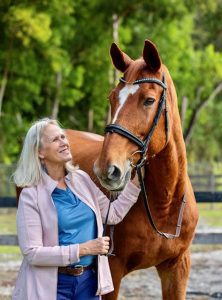 Janet, a Strides member with long blond hair, smiles as she stands outdoors in a light pink jacket and blue shirt, holding the bridle of a tall brown horse with a white blaze. Lush green trees fill the background.