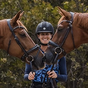 A smiling person in a riding helmet stands between two chestnut horses whose heads are touching, forming a heart shape—capturing true harmony and connection. Lush green foliage fills the background in this V1 Round 2024 moment.