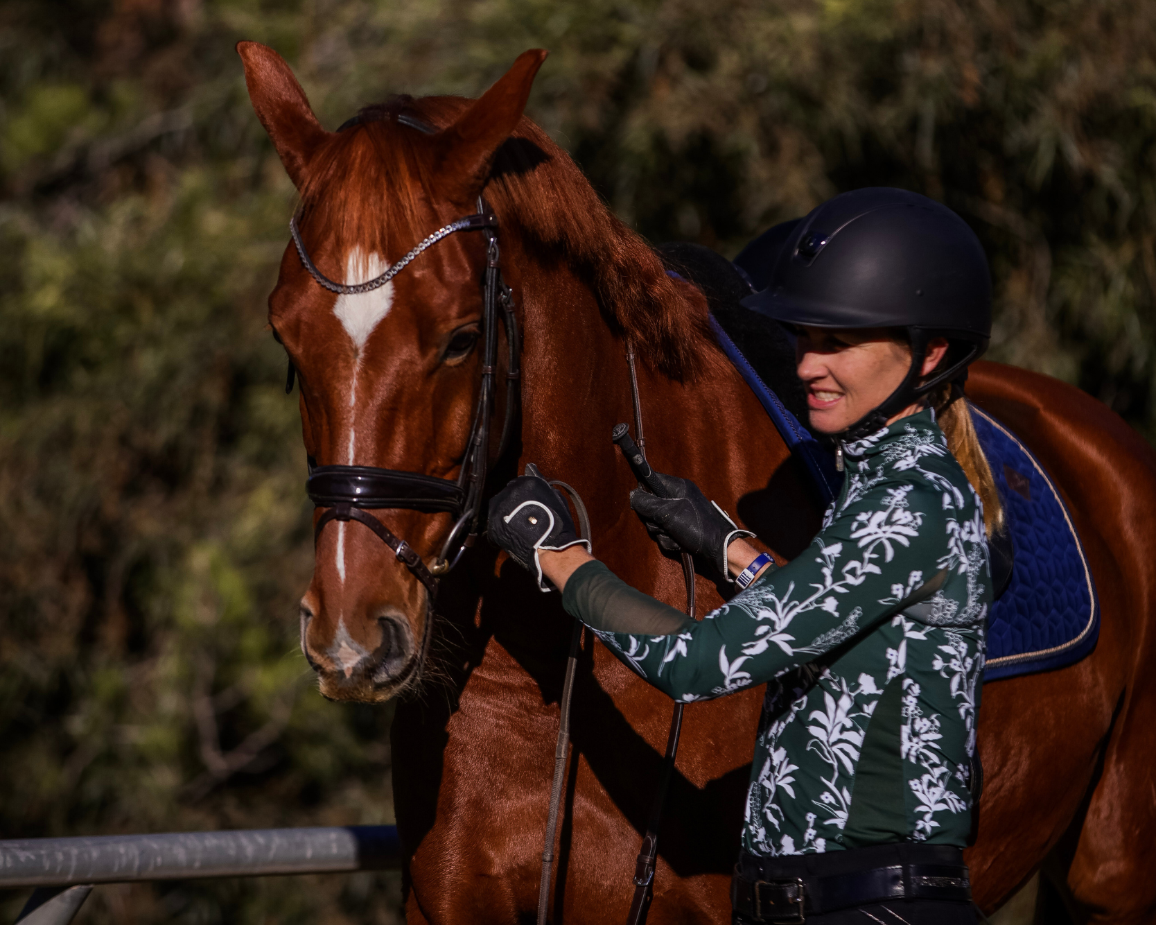 A woman in a dark helmet and patterned shirt stands beside a chestnut horse, holding its bridle and smiling, ready for a round of fitness riding among the trees in the background.