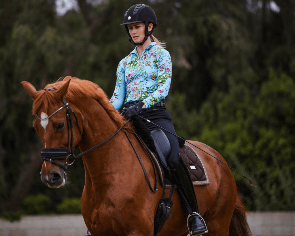 A woman wearing a black helmet and a floral long-sleeve shirt rides a chestnut horse with a white blaze on its face, outdoors amid trees and greenery—an inspiring moment on her 30 Days to Round journey.