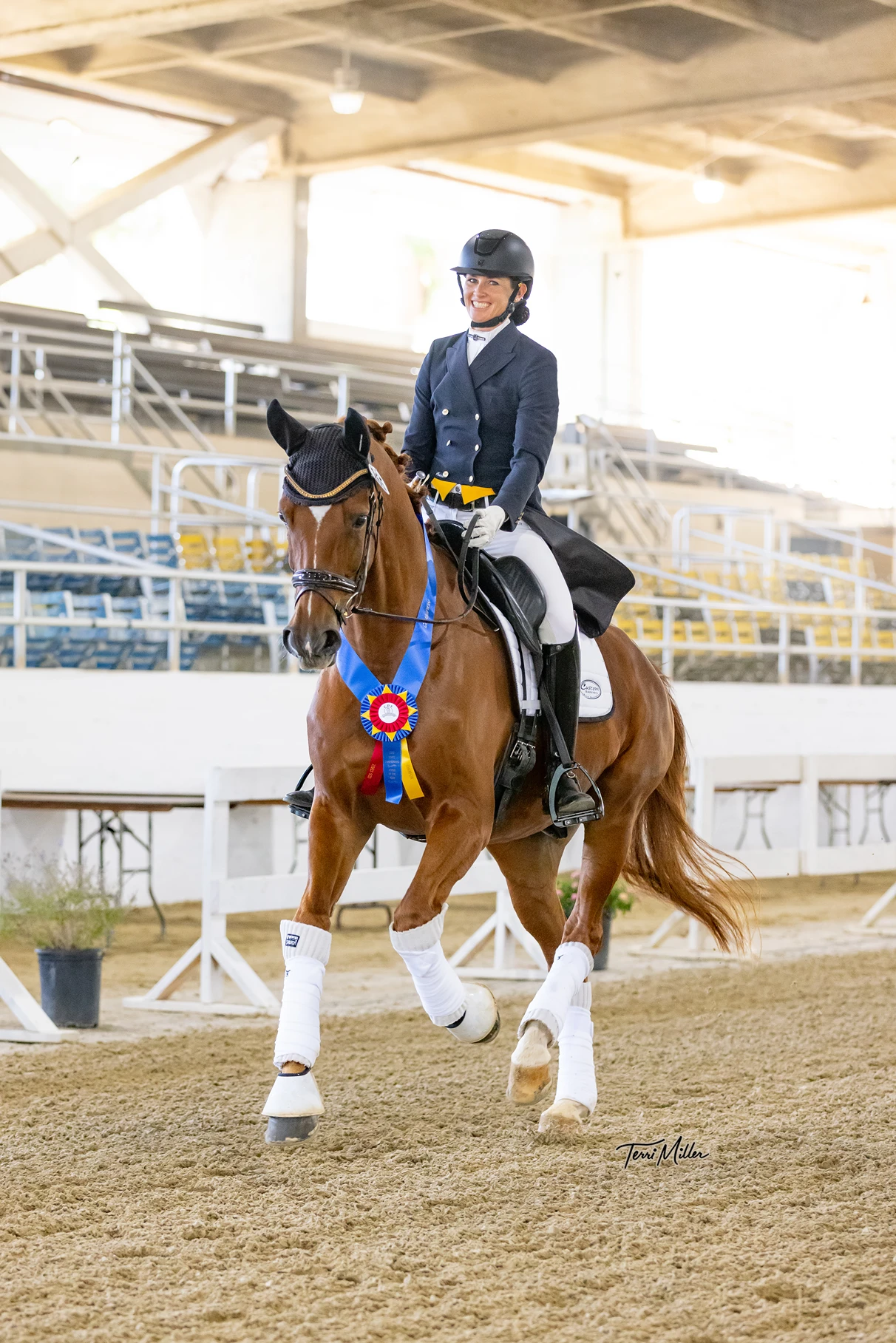 A rider in formal equestrian attire smiles while riding a chestnut horse wearing a blue, yellow, and red rosette. They are indoors, with empty bleachers in the background, on a sandy arena floor.