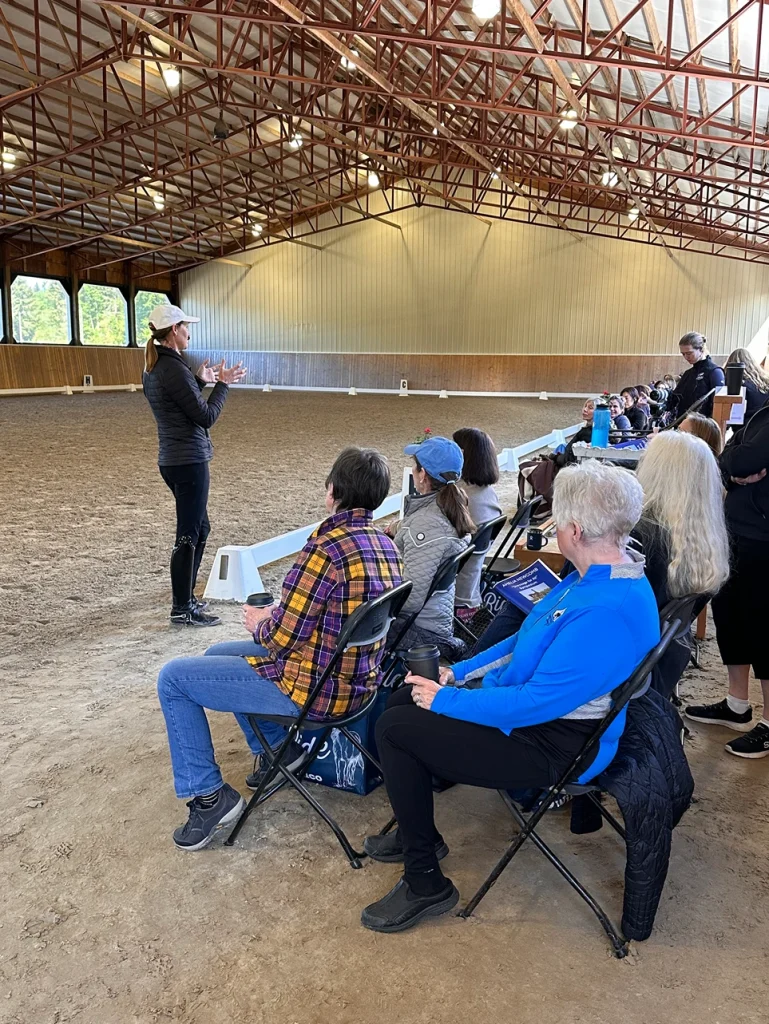 A woman stands and gestures while speaking to a seated audience in an indoor riding arena with a dirt floor and high ceiling. The attendees listen attentively, some taking notes or holding programs.