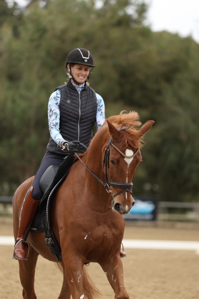 Amelia, smiling in a riding helmet and vest, is enjoying winter riding on a chestnut horse in an outdoor arena, with trees visible in the background.