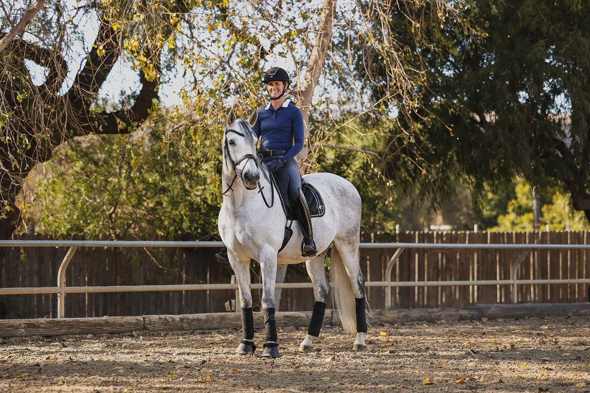 A person wearing a helmet and riding gear sits on a white horse in an outdoor arena, surrounded by a wooden fence and autumn trees—perfect for highlighting the Strides 2024 update on your sales page.