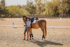 A woman standing and smiling next to a brown horse in an outdoor dressage arena, holding its reins. Both are dressed for riding, ready for dressage training, with trees and riding markers visible in the background.