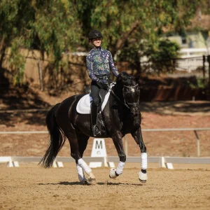 A person wearing a helmet and floral-patterned shirt demonstrates excellent Rider Position while riding a black horse with white leg wraps and a white saddle pad in an outdoor arena, framed by trees and a fence.