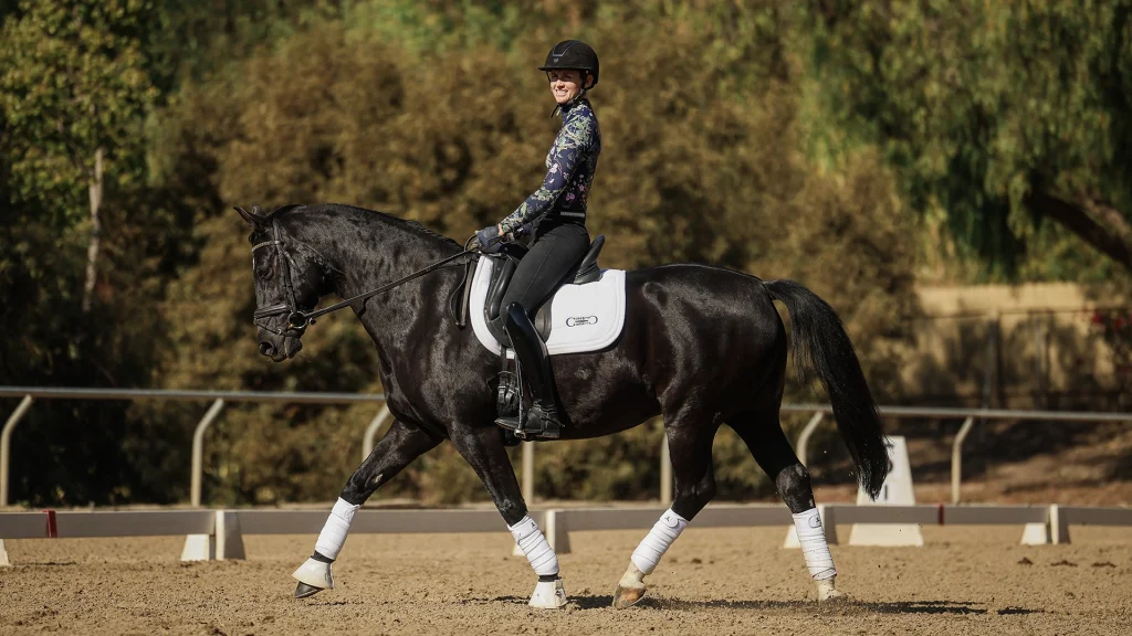 A person in a helmet and riding attire is smiling while showing excellent rider position on a dark horse with white leg wraps and a white saddle pad in an outdoor arena surrounded by trees.