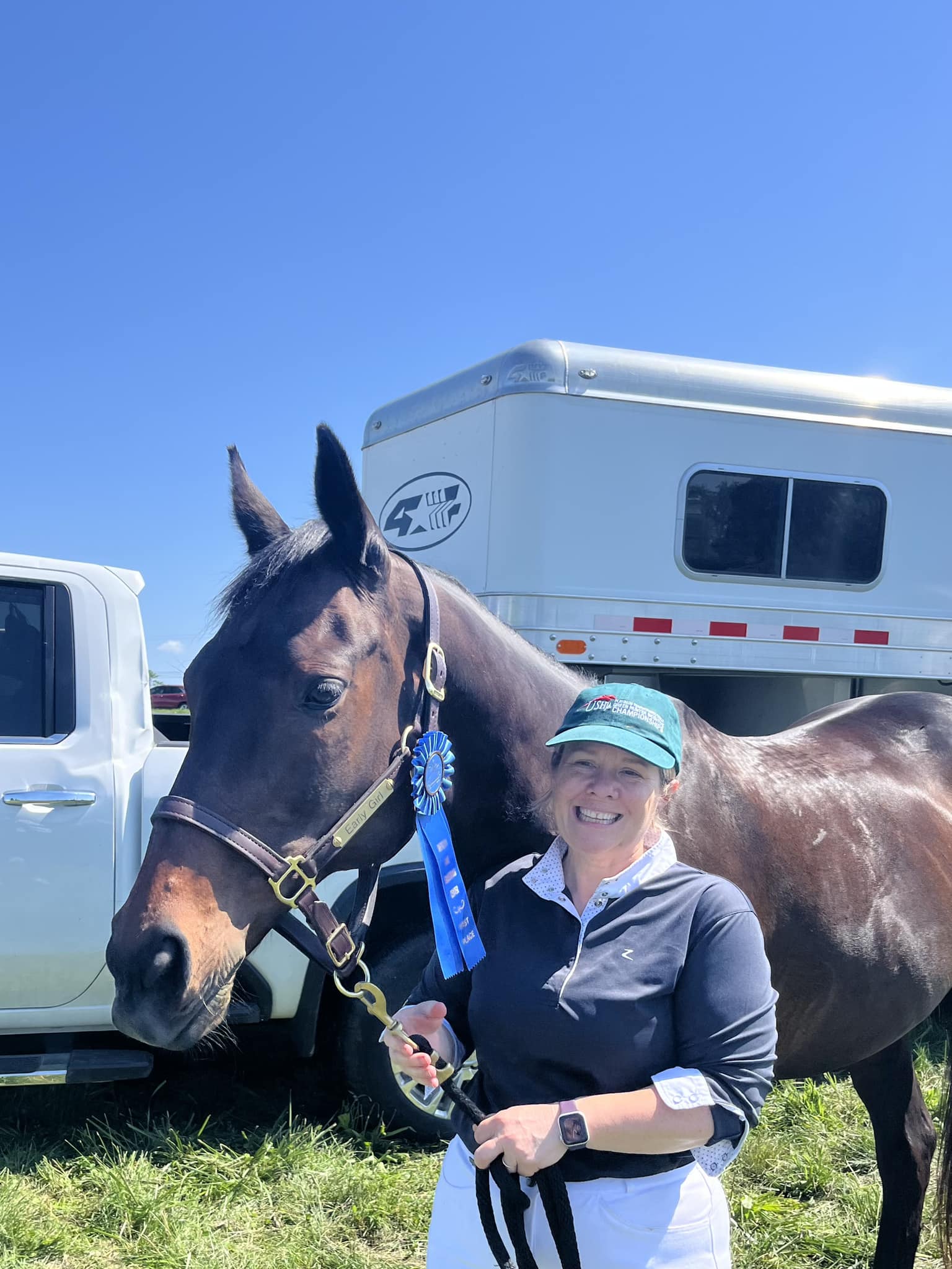 A smiling woman in a green cap holds a dark brown horse with a blue ribbon on its bridle, standing in front of a white truck and horse trailer on a sunny day.