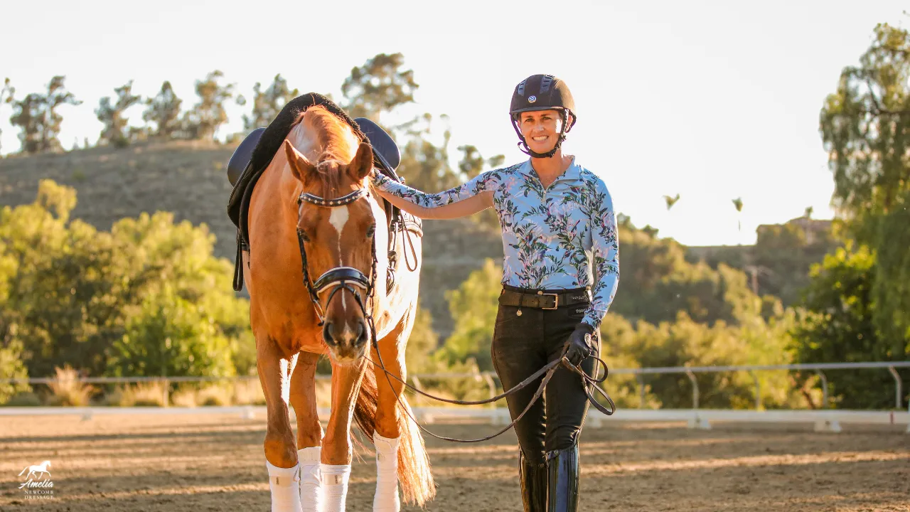 A smiling person in a helmet and riding clothes stands outdoors holding the reins of a brown horse. The fresh air, bright trees, and sunny sky create the perfect backdrop for our Spring newsletter featuring horses in action.