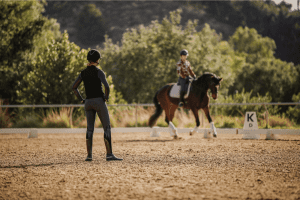A person in riding gear stands on a sandy arena, watching another canter V1 around the ring. Trees and greenery fill the background, with the letter K sign visible—an inviting scene for any Open Page of equestrian life.