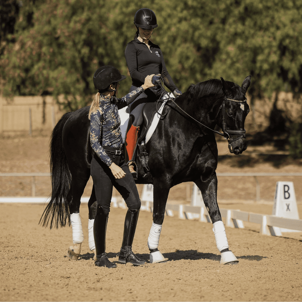 Two women in riding clothes and helmets work with a black horse in an outdoor arena. One woman is mounted, focusing on her rider position, while the other stands beside the horse, offering guidance. White markers and trees are visible in the background.