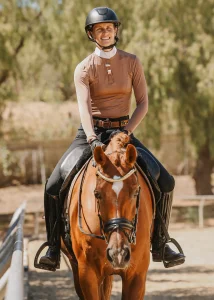 A smiling person wearing a riding helmet and beige long-sleeve shirt sits on a chestnut horse outdoors, preparing for the 2025 Open. Trees and fencing line the background, setting the scene for Oli B’s 30 Days to Round challenge.