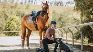 A person wearing an equestrian helmet kneels next to a dog while holding its collar, with a saddled brown horse standing nearby outdoors near a fenced area surrounded by trees at the 2025 Open, part of Oli B’s 30 Days to Round event.