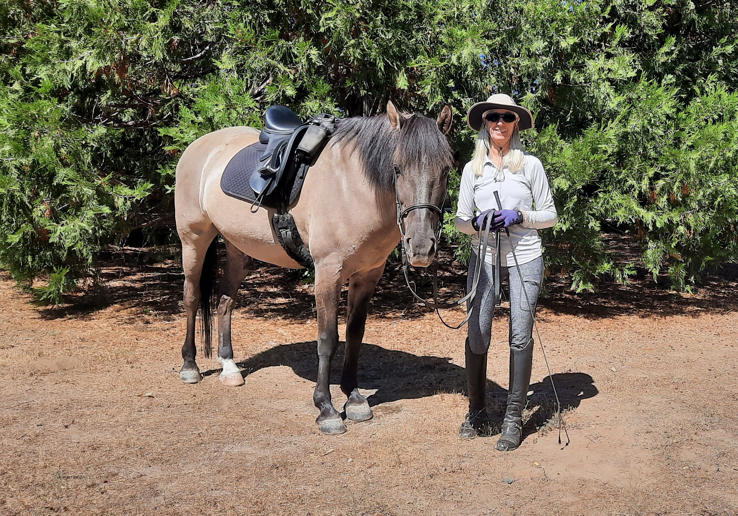 Cindy, a Strides member in riding clothes and sunglasses, stands smiling beside a saddled horse in a sunny, wooded area with green trees in the background.