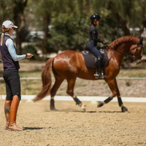 A woman in riding gear instructs another rider on a chestnut horse in an outdoor arena during a 30 Days training program. The coach stands on the sand as the rider and horse move past, with trees and a fence visible in the background.