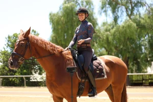 A person wearing a helmet and riding clothes sits smiling on a chestnut horse in an outdoor arena, with trees and clear sky in the background. A PIVO camera is set up on a tripod in front of the horse.