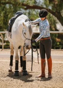 A woman in riding gear smiles and pets a white horse with excellent ground manners, both standing in a sandy outdoor arena on a sunny day.