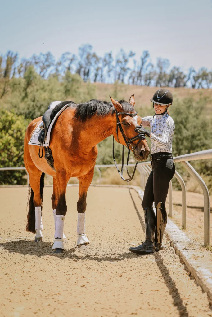 A woman in equestrian gear stands beside a brown horse on a sandy riding arena, smiling and gently petting the horse’s head—showcasing the importance of groundwork essentials. Trees and a blue sky are visible in the background.