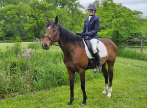 Member Christy, wearing a riding helmet and formal equestrian attire, sits atop a brown horse with a white saddle pad outdoors, enjoying strides across grassy areas with green trees and flowers in the background.