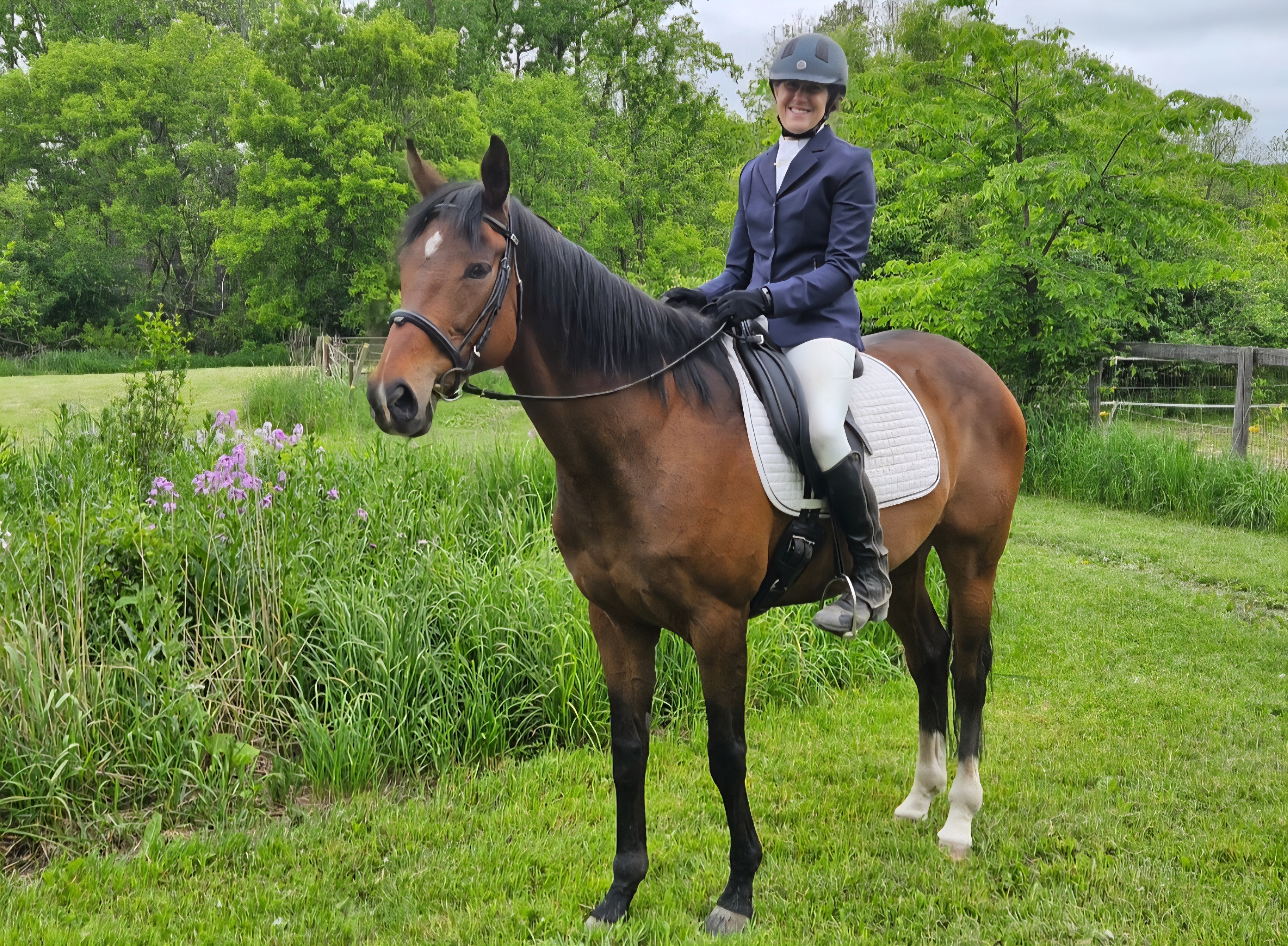 Member Christy, wearing a riding helmet and formal equestrian attire, sits atop a brown horse with a white saddle pad outdoors, enjoying strides across grassy areas with green trees and flowers in the background.