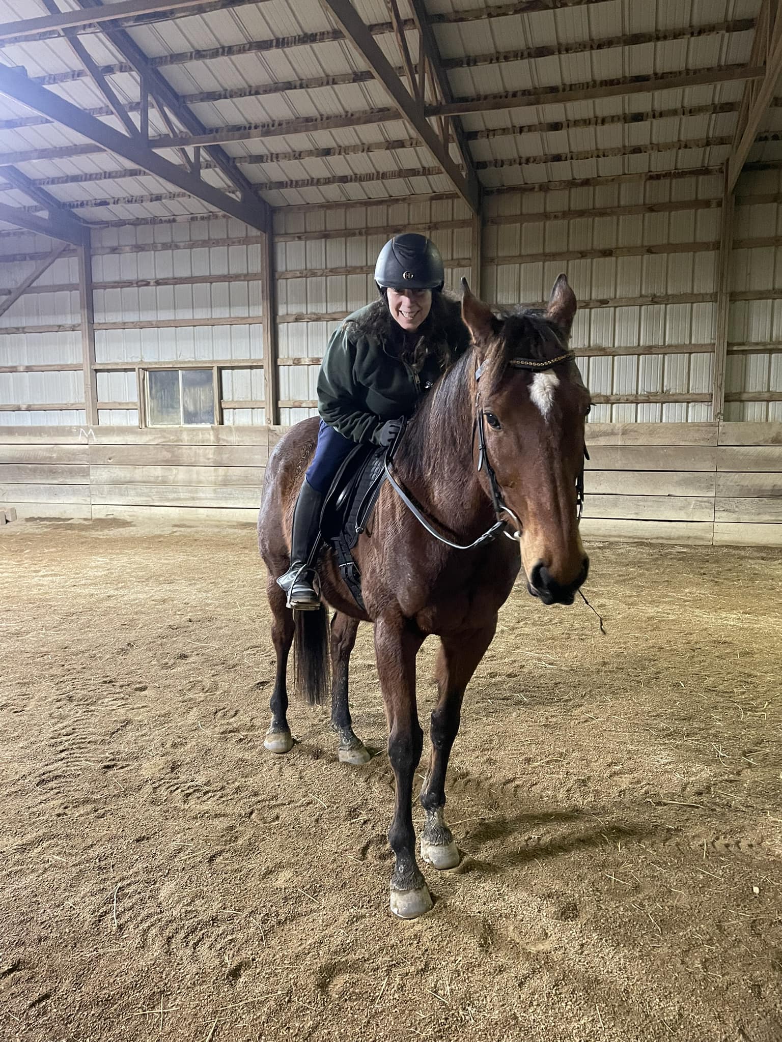 Strides member Wendi, wearing a helmet and dark clothing, is sitting and smiling on a brown horse inside a large indoor riding arena with a dirt floor and wooden walls.