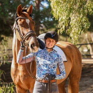 A smiling person in a riding helmet and patterned shirt stands next to a brown horse with a bridle and saddle, holding the reins—celebrating the Grassroots Giving program amid greenery and sunlight in the background.
