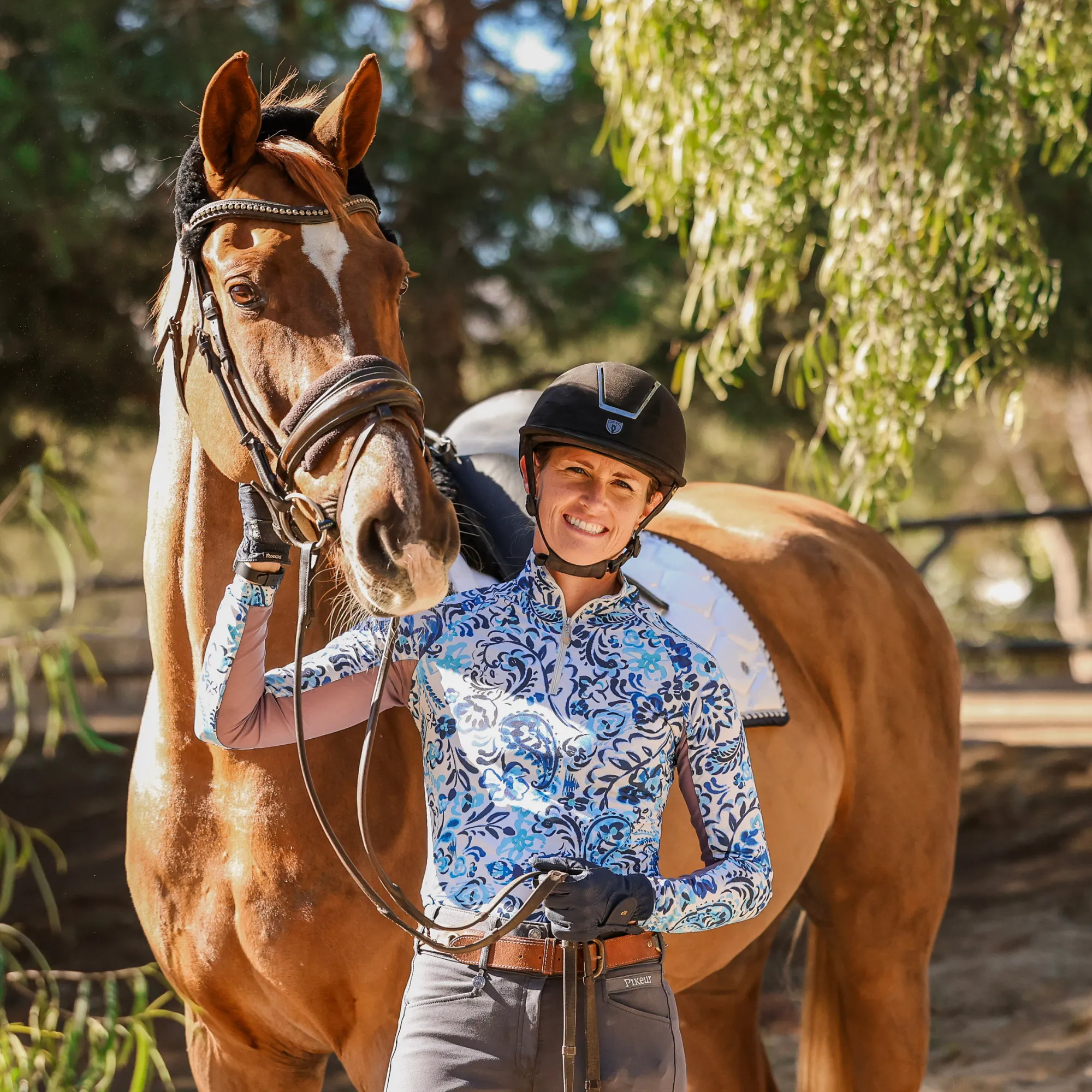 A smiling person in a riding helmet and patterned shirt stands next to a brown horse with a bridle and saddle, holding the reins—celebrating the Grassroots Giving program amid greenery and sunlight in the background.
