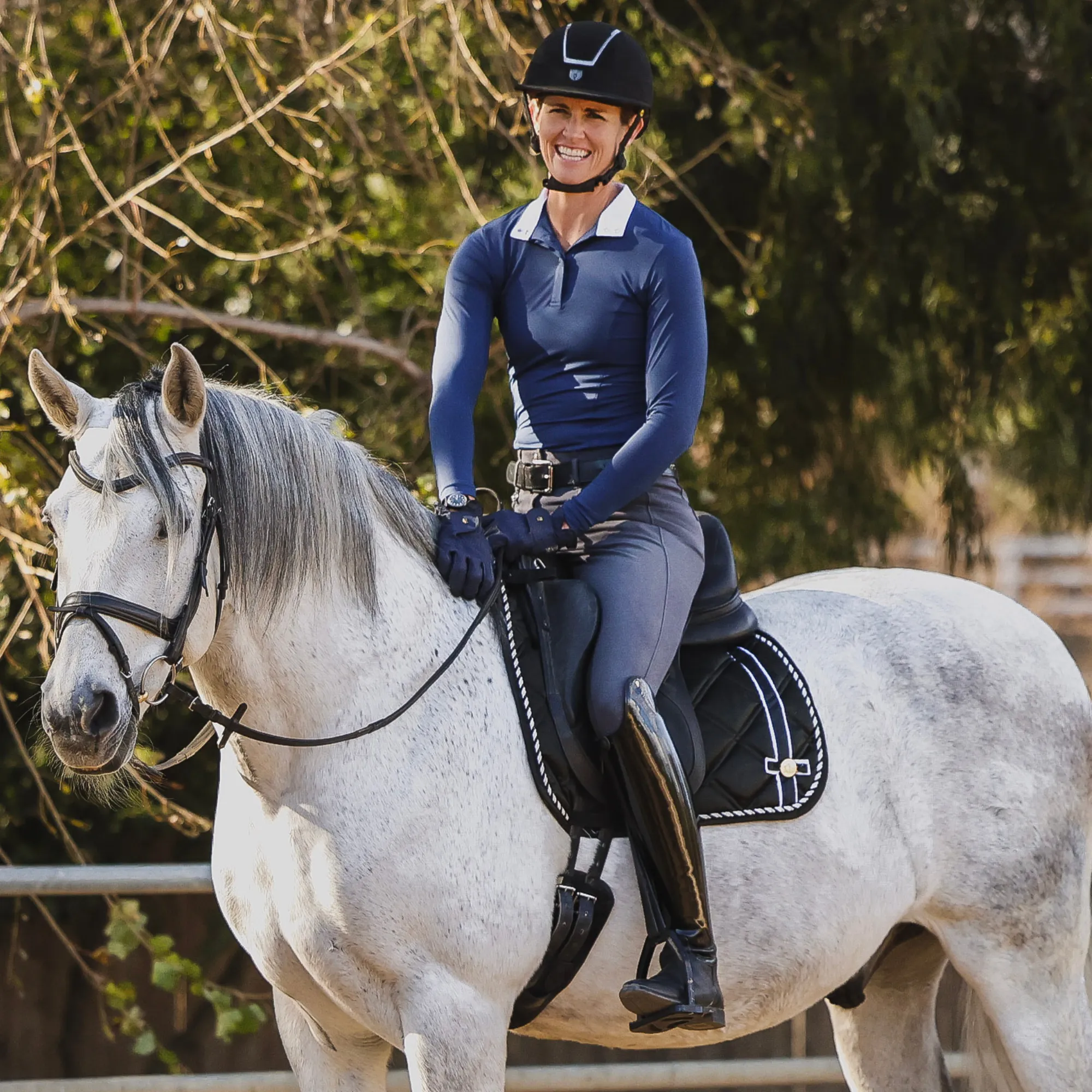 A person in equestrian gear, including a helmet and boots, is smiling while riding a white horse outdoors near trees and a wooden fence during a Grassroots riding program.