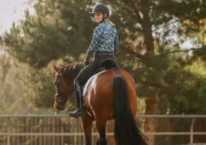 A person wearing a helmet and patterned shirt rides a brown horse outdoors near trees, looking back and smiling, as they enjoy practicing dressage.